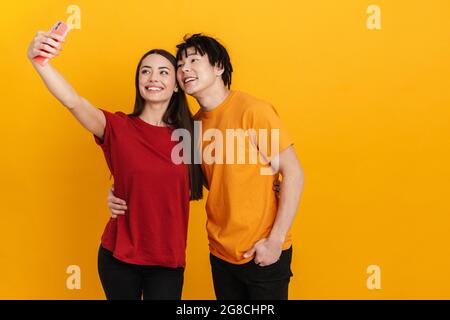 Sorridente coppia multirazziale abbracciando mentre prendendo selfie sul cellulare isolato sopra parete gialla Foto Stock