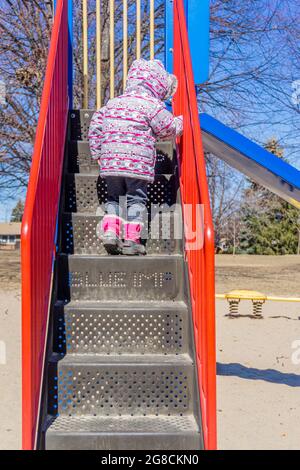 Toronto, Canada, marzo 2018 - Vista posteriore del bambino con giacca invernale e stivali sulle scale di un'attrezzatura scorrevole su un parco giochi per bambini Foto Stock