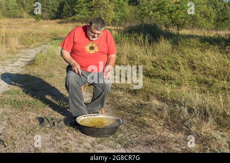 Vecchio divertente uomo caucasico anziano pesca piccolo pesce in bacino antico mentre si siede su uno sgabello Foto Stock