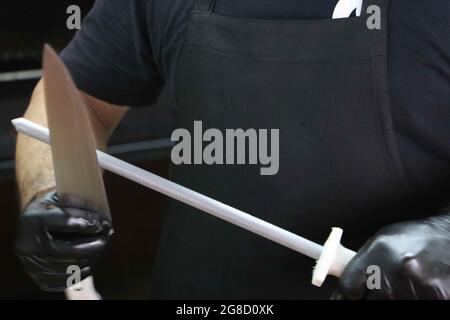 uomo affilando un coltello da barbecue sulla seggiovia. Con grembiule nero sullo sfondo. Foto Stock