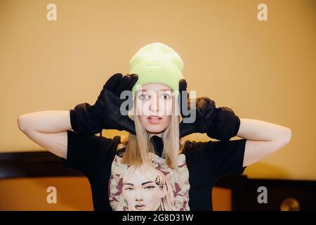 Bella donna bionda che indossa un mix di abiti invernali ed estivi al chiuso, cappello, guanti, t-shirt a maniche corte, ci guarda con le mani accanto alla sua testa Foto Stock