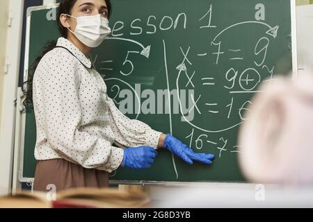 Insegnante femminile che indossa una maschera facciale con guanti in lattice e che spiega una formula matematica alla classe Foto Stock