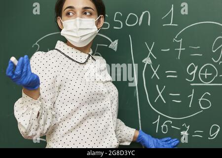 Insegnante femminile che indossa una maschera facciale con guanti in lattice e che spiega una formula matematica alla classe Foto Stock