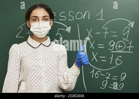 Insegnante femminile che indossa una maschera facciale con guanti in lattice e che spiega una formula matematica alla classe Foto Stock
