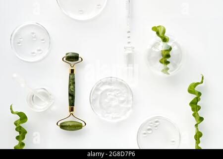 Moisturizer e rullo per il viso in giada verde. Foglie esotiche di felce e gocce d'acqua su sfondo bianco. Massaggio viso, cosmetici fatti a mano. Foto Stock