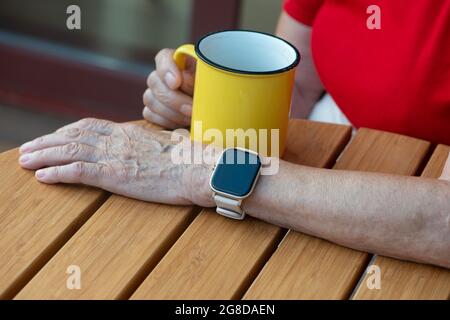 Donna anziana con moderno orologio intelligente bere caffè del mattino. Orologio intelligente con schermo nero vuoto, spazio per il tuo design. Assistenza sanitaria e tecnologia co Foto Stock