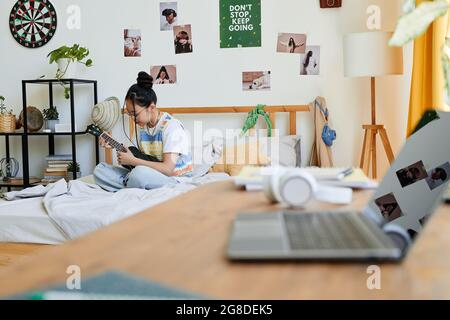 Ritratto di ragazza teenage asiatica che gioca ukulele mentre si siede sul letto in una stanza accogliente, spazio copia Foto Stock