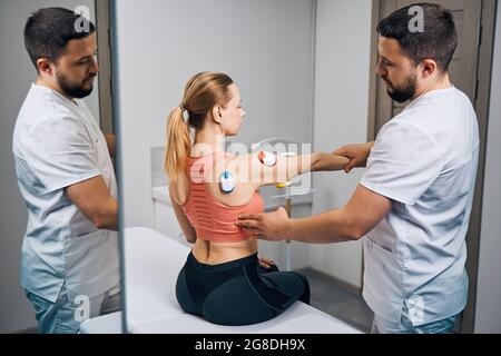 Miostimolazione degli elettrodi per terapia fisica. Dottore in uniforme mette l'elettrostimolatore sulla mano della donna Foto Stock