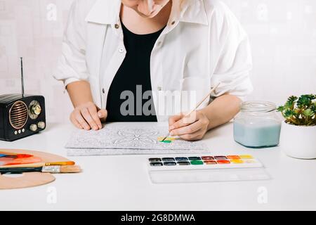 Giovane donna colorazione pagina antistress a tavola al chiuso, benessere mentale e arte terapia. Donna dipinge uno schizzo, meditativo processo di colorazione pagine. Foto Stock