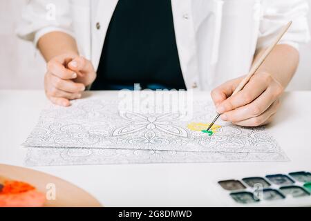 Giovane donna colorazione pagina antistress a tavola al chiuso, benessere mentale e arte terapia. Donna dipinge uno schizzo, meditativo processo di colorazione pagine. Foto Stock