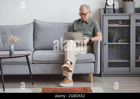 Ritratto a tutta lunghezza di un uomo anziano moderno che usa un computer portatile a casa mentre si siede sul divano in un ambiente interno minimo, spazio per la copia Foto Stock