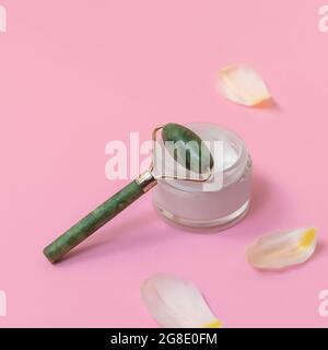 Massaggio viso Jade Roller con crema viso su rosa pastello sfondo con petali bianchi Foto Stock