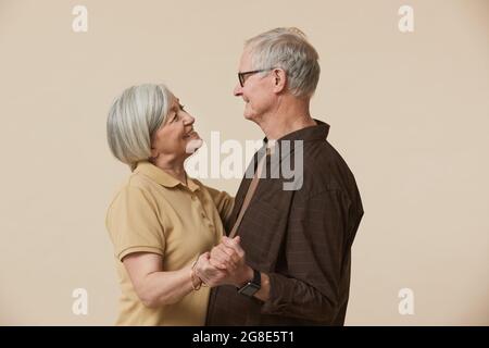 Ritratto minimale in vita di coppia senior felice che danzano e si guardano l'un l'altro su sfondo beige Foto Stock