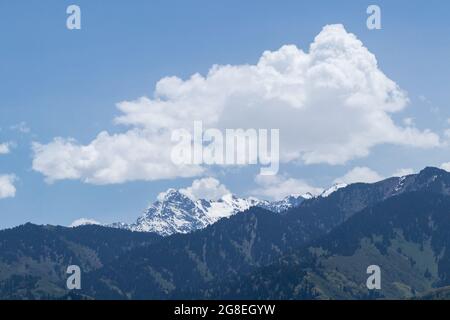 Una vetta innevata dietro le colline e le nuvole bianche sopra di essa. Contro il cielo blu Foto Stock