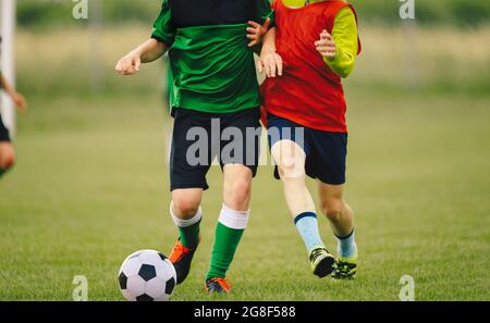 Due giocatori di calcio in un Duel on Grass Venue. Ragazzi che corrono dopo la palla da calcio bianca e nera. Kids in divise e scarpe da calcio verdi e rosse Foto Stock