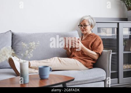 Ritratto a lunghezza intera della donna anziana moderna che usa lo smartphone mentre si sdraiava sul divano in un ambiente domestico minimo, spazio di copia Foto Stock