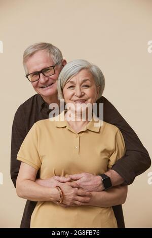 Ritratto minimale della coppia senior che abbraccia e guarda la fotocamera mentre si sta in piedi su sfondo beige Foto Stock
