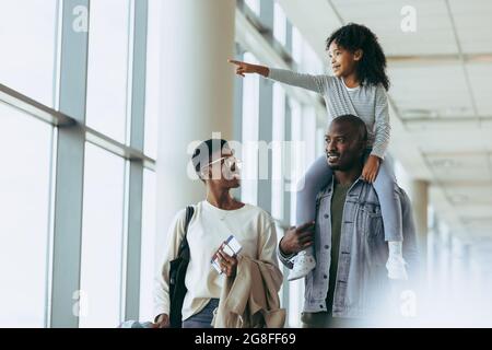 Felice famiglia africana che va in vacanza in aeroporto. Coppia che cammina in aeroporto con la figlia che si siede a spalla e che punta via. Foto Stock