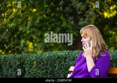 Defocus bionda caucasica donna che parla, parlando al telefono esterno, all'aperto. donna di 40 anni in blusa viola in panchina. Persona adulta usi Foto Stock