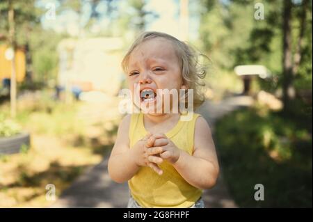 Primo piano ritratto di piccolo divertente bambina bionda bambina in tuta gialla piangendo all'esterno in estate. Tantrum infantile. Concetto sano di infanzia Foto Stock