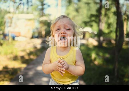 Primo piano ritratto di piccolo divertente bambina bionda bambina in tuta gialla piangendo all'esterno in estate. Tantrum infantile. Concetto sano di infanzia Foto Stock