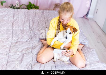 Una ragazza adolescente con un telefono cellulare, abbracciando un cane Jack Russell Terrier, comunica sui social network o via Zoom con amici o familiari, fa un Foto Stock