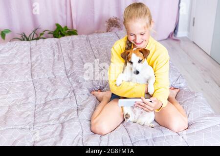 Una ragazza sorridente con un telefono cellulare, abbracciando un cane Jack Russell Terrier, comunica sui social network con gli amici o via Zoom, effettua una videochiamata Foto Stock