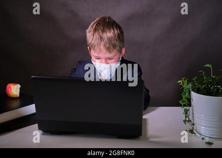 Un ragazzo in una tuta da scuola con una maschera medica sul viso a un computer, copiare spazio su uno sfondo scuro studio Foto Stock