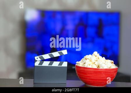 Clapper di film nero e una ciotola di popcorn, televisione sfocata in background. Foto di sfondo del settore dell'intrattenimento. Tempo filmato. Guardare la tv. Foto Stock