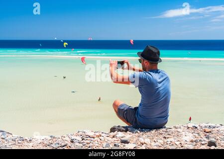 Turista con cappello di paglia che fotografa kiteboarders con smartphone alla spiaggia di Sotavento, Jandia, Fuerteventura, Isole Canarie, Spagna, Atlantico, Europa Foto Stock