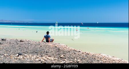 Uomo seduto su rocce che guardano i kitesurfers nel mare limpido, spiaggia di Sotavento, Jandia, Fuerteventura, Isole Canarie, Spagna, Atlantico, Europa Foto Stock