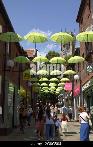 Una vista lungo l'area pedonale di Copper Gate a york, Umbrellee colorate di Lime pendono tra gli edifici e la torre di tutti i Santi Chiesa può essere Foto Stock