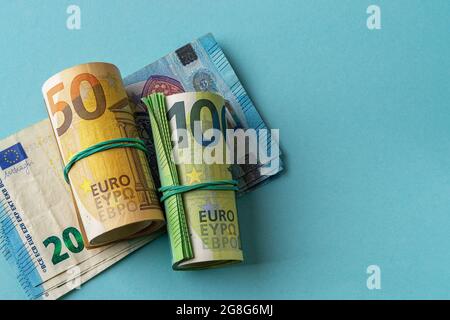 Rotolo verde di banconote da 100 euro e rotolo arancione di banconote da 50 euro oltre 20 euro di carta moneta su sfondo blu. Reddito, spese e risparmio in contanti. Foto Stock