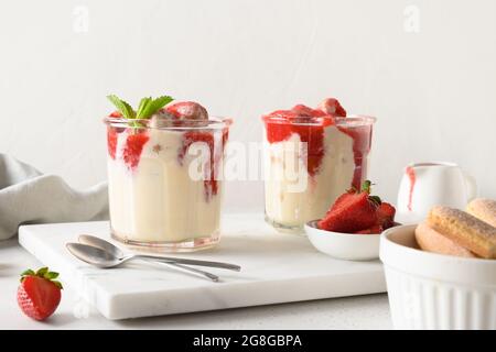 Dessert a strati in vasetti di vetro con biscotti savoyardi, mascarpone e fragole decorate con panna montata. Tiramisù italiano con vibrazioni estive su bianco Foto Stock