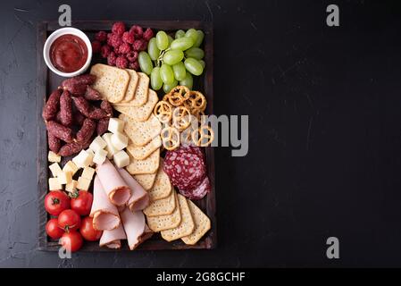 Pomodori, salsicce, frutta, cracker e formaggio su salumi a bordo su sfondo nero, spuntini da festa, primo piano. Foto Stock
