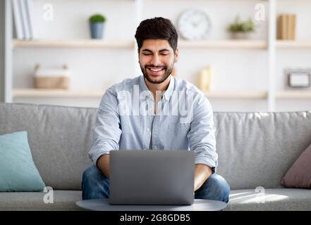 Concetto di lavoro freelance. Allegro uomo d'affari arabo che lavora su un computer portatile online e sorridente seduto sul divano a casa Foto Stock