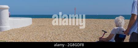 Una vista laterale e posteriore di un anziano sconosciuto in una sedia a rotelle con assistente, guardando verso una spiaggia Pebble e vista mare. Foto Stock