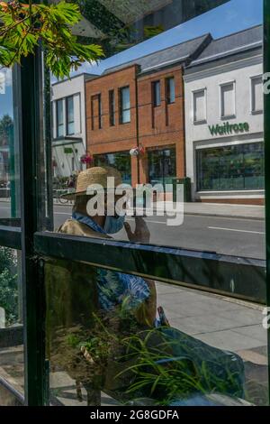 Dorking, Surrey, UK, 07-19-2021: Una persona sconosciuta seduto con il cappello di paglia una maschera facciale e fumare un sigaro ad una fermata dell'autobus, fuori del Waitrose locale. Foto Stock