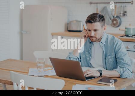 Uomo d'affari serio messo a fuoco bearded in vestiti casual che lavorano sul portatile mentre si siede al tavolo della cucina Foto Stock