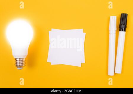 Lampada a risparmio energetico incandescente con carta bianca per appunti poggiata su un tavolo giallo, modello vuoto con spazio di copia - Vista dall'alto, disposizione piatta Foto Stock
