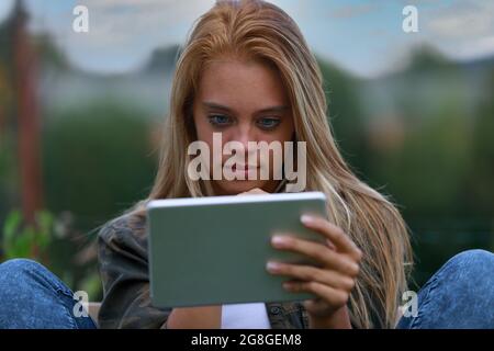 giovane donna seduta informale su una panchina mentre si gode di contenuti digitali, forse in social network, navigando in internet o video Foto Stock