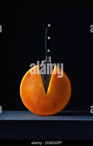 Grande tondo di formaggio duro con coltello sulla scrivania in ardesia su sfondo scuro. Foto Stock