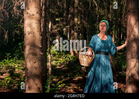 giovane donna in abiti popolari contadini con un cesto di vimini alcune piante selvatiche, bacche o funghi nella foresta di conifere Foto Stock