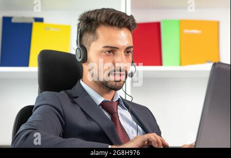 Ritratto di un uomo d'affari, un addetto del call center che indossa cuffie con microfono e lavora con un computer portatile in ufficio. Uomo in tuta consulente cliente su telefono in personalizzato Foto Stock