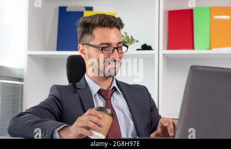 Ritratto di felice uomo d'affari in abbigliamento formale che lavora con il computer portatile in ufficio. Uomo allegro e rilassato in tuta che lavora mentre beve caffè o tè. Foto Stock
