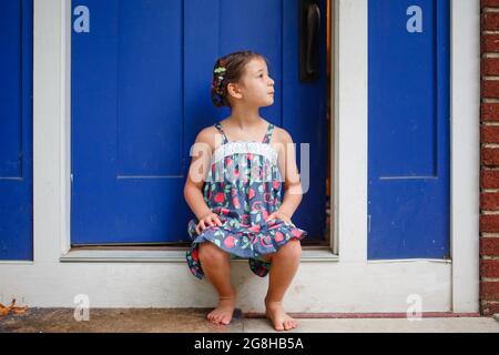 Carino ragazza piccola con trecce siede a piedi nudi in Sundress sul fronte stoop Foto Stock