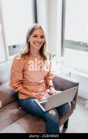 Una donna sorridente si siede sul divano dell'ufficio mentre lavora sul laptop Foto Stock