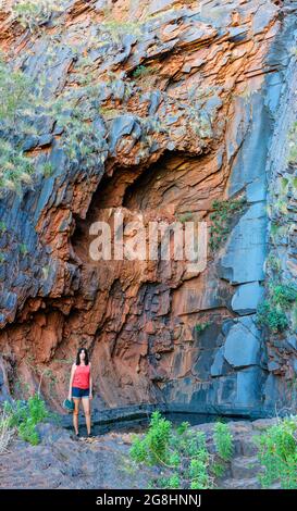 Una donna che cammina nel bush si trova accanto alla piscina ammirando la parete rocciosa verticale, asciutta ma impressionante, delle cascate Perentie sulla stazione di Cheela, Australia Occidentale. Foto Stock