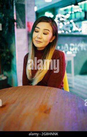 Donna asiatica con gli occhi chiusi seduti e in posa al tavolo nel caffè. Bella ragazza in borgogna turtleneck siede nel ristorante. Illumina un contatore a barre sullo sfondo. Riposo, divertimento per il tempo libero. Foto Stock