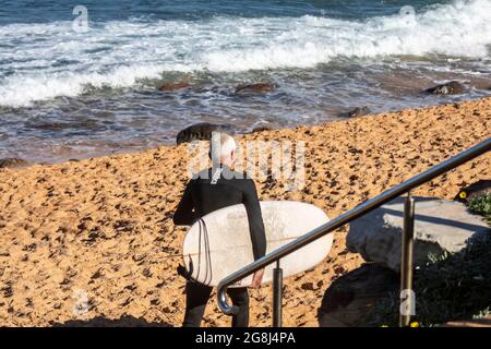 Surfer australiano di media età che porta la sua tavola da surf all'oceano, Sydney, Australia Foto Stock